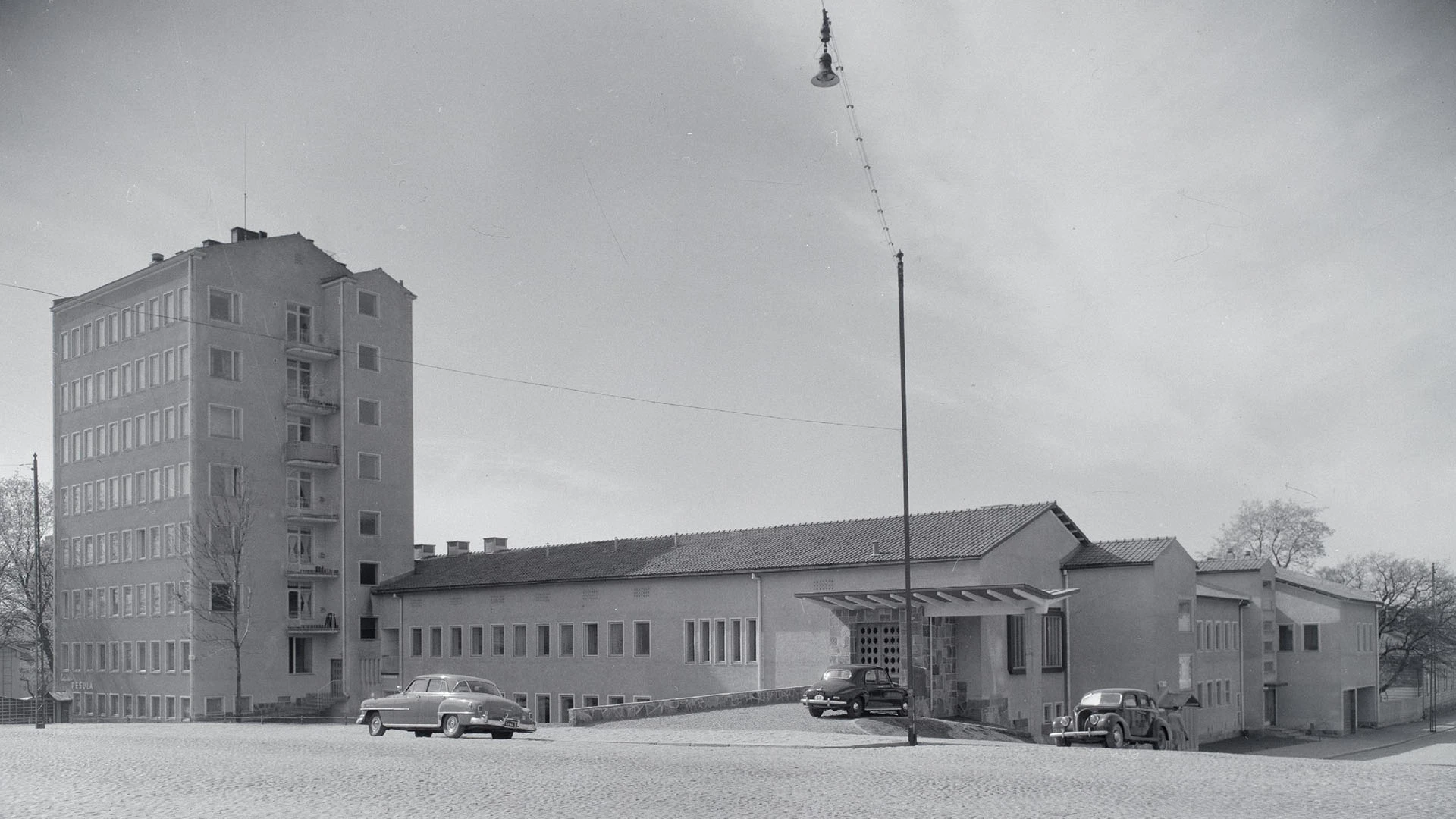 Vanha valokuva sairaalarakennuksesta, etualalla 1950-luvun autoja.